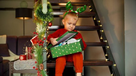 Foto de Lindo niño abre la caja de regalo de Navidad y se emociona de sus nuevos juguetes. Celebraciones familiares en las vacaciones de invierno - Imagen libre de derechos
