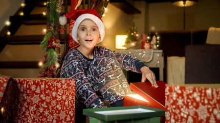 Foto de Retrato de niño sonriente emocionado en pijama abriendo regalos de Navidad y regalos de Santa por la noche en la sala de estar. - Imagen libre de derechos