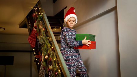 Foto de Niño pequeño en pijama bajando las escaleras sosteniendo una pila de cajas de regalo de Navidad con regalos. Celebraciones familiares en las vacaciones de invierno. - Imagen libre de derechos