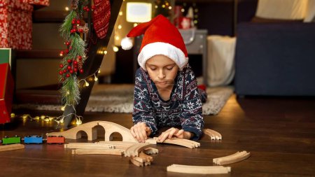 Téléchargez les photos : Garçon mignon construisant chemin de fer jouet qu'il a reçu du Père Noël comme cadeau de Noël ou du Nouvel An. Célébrations familiales pendant les vacances d'hiver. - en image libre de droit