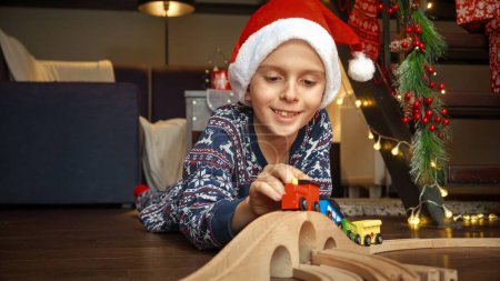 Téléchargez les photos : Portrait d'un petit garçon heureux célébrant Noël couché sur le sol et jouant avec un train en bois coloré et un chemin de fer en bois. Vacances d'hiver, fêtes et fêtes - en image libre de droit