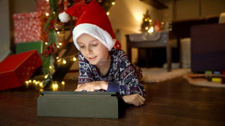 Téléchargez les photos : Joyeux garçon souriant portant le chapeau du Père Noël couché sur le sol dans le salon et jouant sur tablette. Vacances d'hiver, fêtes et fêtes - en image libre de droit