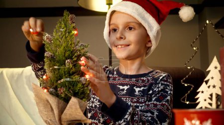 Foto de Retrato del niño sonriente en el sombrero de Papá Noel decorando un pequeño árbol de Navidad con luces coloridas y guirnaldas. Vacaciones de invierno, celebraciones y fiestas - Imagen libre de derechos
