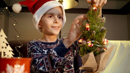 Foto de Feliz niño sonriente disfrutando de la decoración del árbol de Navidad con luces y guirnaldas en casa. Vacaciones de invierno, celebraciones y fiestas - Imagen libre de derechos