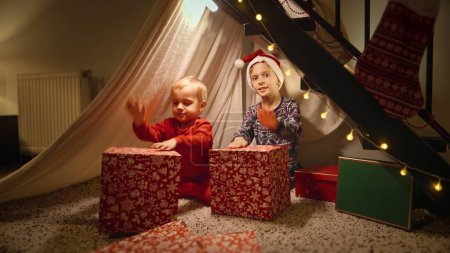 Téléchargez les photos : Deux garçons souriants heureux célébrant Noël assis dans une tente de tipi et jouant avec des cadeaux et des cadeaux de Noël. Vacances d'hiver, fêtes et fêtes - en image libre de droit