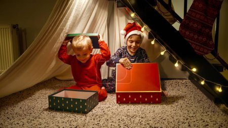 Photo for Two cute little boys in pajamas sitting under stairs at house and looking at Christmas presents from Santa - Royalty Free Image