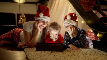 Photo for Happy family celebrating Christmas lying on floor and watching video on tablet computer in living room. Winter holidays, celebrations and party - Royalty Free Image