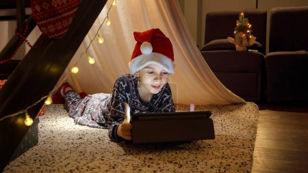 Foto de Retrato de niño alegre esperando a Santa en Navidad jugando juegos en la tableta. Vacaciones de invierno, celebraciones y fiestas - Imagen libre de derechos