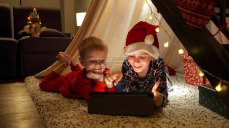Téléchargez les photos : Deux petits garçons ayant une conversation vidéo sur tablette alors qu'ils étaient couchés sur le sol dans le salon décoré pour Noël. Parler à leur grand-mère pendant les vacances d'hiver - en image libre de droit