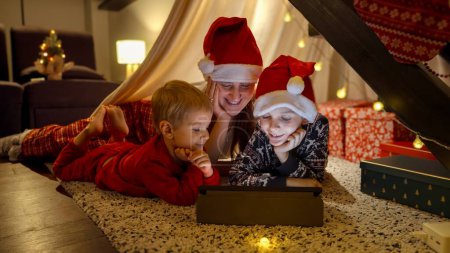 Photo for Happy mother with two sons lying on floor and using tablet computer on a Christmas night. Winter holidays, celebrations and party - Royalty Free Image