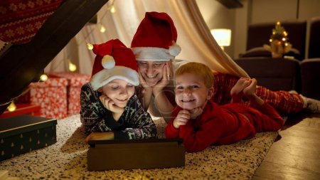 Photo for Two happy boys with mother celebrating Christmas and watching movie or cartoon on tablet computer while playing in tepee tent. Winter holidays, celebrations and party - Royalty Free Image