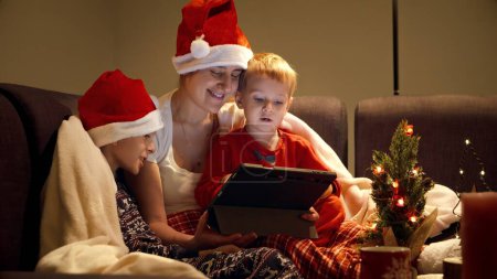 Téléchargez les photos : Mère heureuse avec des enfants regardant le film de Noël traditionnel sur tablette tout en se relaxant sur le canapé dans le salon. Vacances d'hiver, fêtes et fêtes - en image libre de droit