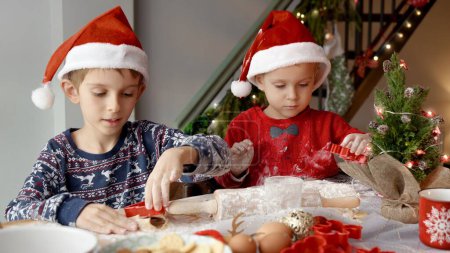 Téléchargez les photos : Portrait de deux petits garçons en chapeaux SAnta appréciant rouler la pâte et découper les biscuits de Noël et le pain d'épice pour les célébrations du Nouvel An. - en image libre de droit
