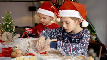 Téléchargez les photos : Bébé garçon avec frère aîné faire de la pâte et découper les biscuits de Noël traditionnels tout en se préparant pour les célébrations du Nouvel An. Vacances d'hiver, fêtes et fêtes - en image libre de droit