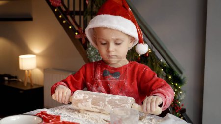 Photo for Adorable toddler boy rolling dough with wooden rolling pin while preparing cookies and biscuits for Christmas. Winter holidays, celebrations and party - Royalty Free Image