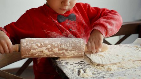 Téléchargez les photos : Gros plan de petit garçon préparant la pâte et utilisant le rouleau à pâtisserie sur la table de cuisine. Vacances d'hiver, fêtes et fêtes - en image libre de droit