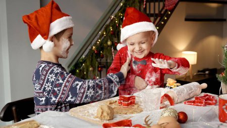 Foto de Dos niño sonriente alegre en los sombreros de Santa divertirse mientras se cocina y tirar harina el uno al otro mientras se prepara para la Navidad. - Imagen libre de derechos