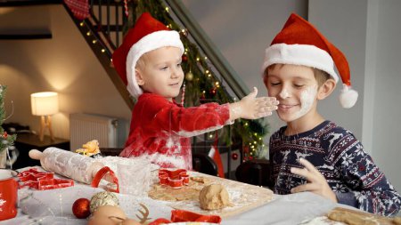 Téléchargez les photos : Deux garçons joyeux jouant et s'amusant avec de la farine tout en cuisinant à Noël à la maison. Vacances d'hiver, fêtes et fêtes - en image libre de droit