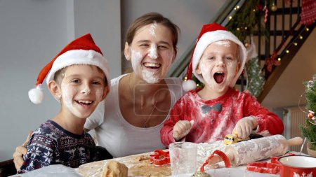 Foto de Niños divertidos felices con la madre sonriendo y riendo en la cámara mientras se ensucia y ensucia durante la cocina y se prepara para la Navidad y el Año Nuevo - Imagen libre de derechos