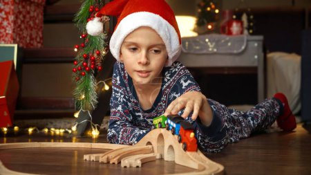 Téléchargez les photos : Joyeux garçon souriant dans le chapeau du Père Noël couché o plancher et jouer avec jouet train en bois et chemin de fer. Vacances d'hiver, fêtes et fêtes - en image libre de droit