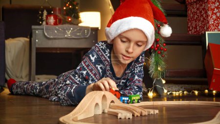 Foto de Niño en pijama jugando con ferrocarril de madera y tren que recibió como regalo en Navidad de Santa. Vacaciones de invierno, celebraciones y fiestas - Imagen libre de derechos