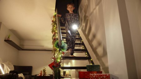 Téléchargez les photos : Mignon petit garçon en pyjama marchant dans les escaliers en bois avec lampe de poche pour chercher des cadeaux de Noël et des cadeaux du Père Noël - en image libre de droit