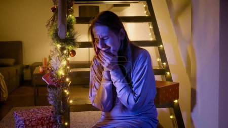 Photo for Police or ambulance car lights illuminating face of crying young woman sitting on wooden stairs decorated for Christmas or New Year. Crime, ambulance, injury, problems with law and theft on winter holidays and celebrations - Royalty Free Image