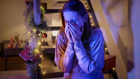 Photo for Young crying woman sitting in living room decorated for Christmas or New Year and looking at police lights and sirens. Crime, ambulance, injury, problems with law and theft on winter holidays and celebrations - Royalty Free Image