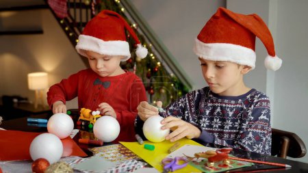 Photo for Two boys in Santa's caps making traditional handmade Christmas decorations and baubles for winter holidays. - Royalty Free Image