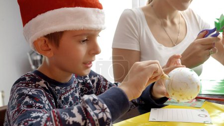Téléchargez les photos : Portrait de petit garçon faisant boule de sapin de Noël à la main et le décorer avec des paillettes. Vacances d'hiver, temps en famille ensemble, enfants avec les parents célébrant - en image libre de droit