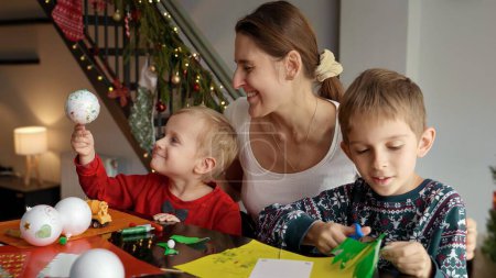 Foto de Feliz madre amorosa mirando a sus hijos haciendo decoraciones navideñas hechas a mano. Vacaciones de invierno, tiempo en familia juntos, niños con padres celebrando - Imagen libre de derechos
