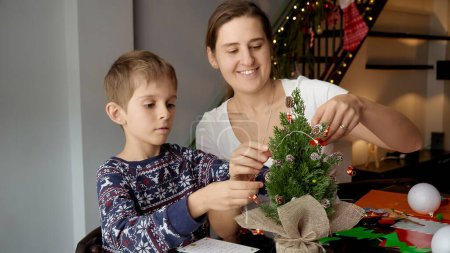 Foto de Niño feliz con madre decorando un pequeño árbol de Navidad con guirnaldas y luces. Vacaciones de invierno, tiempo en familia juntos, niños con padres celebrando - Imagen libre de derechos