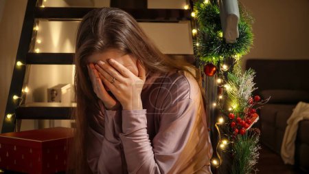 Téléchargez les photos : Jeune femme bouleversée pleurant sur des escaliers en bois laissés seuls à Noël. Émotions négatives, dépression et solitude lors des célébrations - en image libre de droit