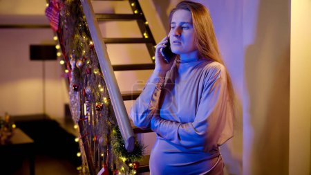 Photo for Portrait of nervous woman talking to emergency service on Christmas night. Police car lights illuminate the room. Crime, ambulance, injury, problems with law and theft on winter holidays and celebrations - Royalty Free Image