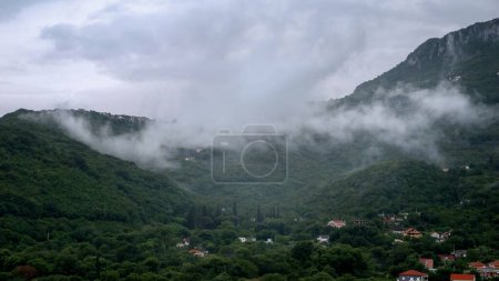 Téléchargez les photos : Petit village en haute montagne recouvert d'arbres et couvert de nuages de faible pluie. - en image libre de droit