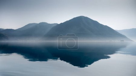 Téléchargez les photos : Mer calme ou vagues de lac, brouillard matinal levant et hautes montagnes bleues au bord de la mer. - en image libre de droit
