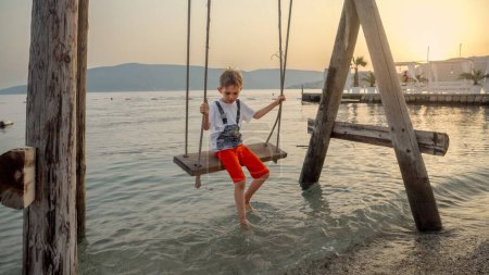Foto de Niño pequeño balanceándose en cuerda de madera columpiarse en la playa al atardecer. Vacaciones, vacaciones de verano y turismo. - Imagen libre de derechos