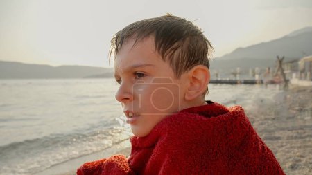 Téléchargez les photos : Portrait de petit garçon se sentant froid après avoir nagé dans la mer et enveloppé dans une serviette rouge. Vacances, vacances d'été et tourisme. - en image libre de droit