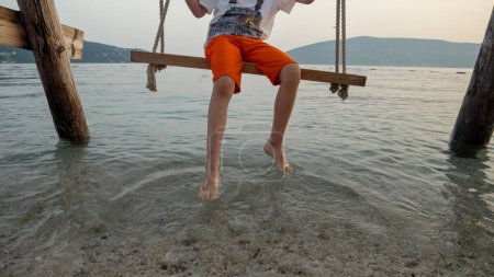 Téléchargez les photos : Gros plan de petit garçon se balançant sur la plage de la mer et touchant les vagues de l'océan avec les pieds. Vacances, vacances d'été et tourisme. - en image libre de droit
