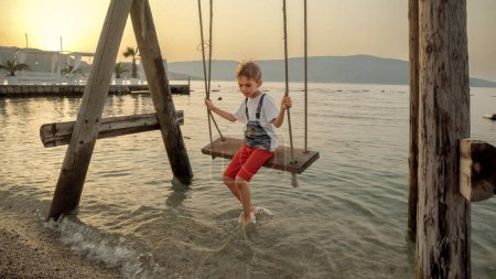 Téléchargez les photos : Petit garçon appréciant monter sur la balançoire à la plage de la mer. Vacances, vacances d'été et tourisme. - en image libre de droit