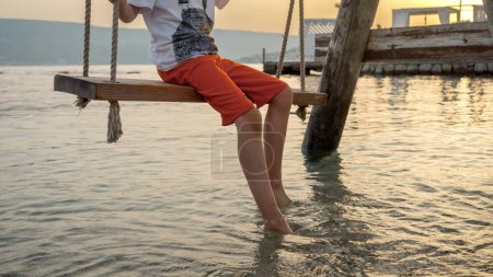 Foto de Primer plano de los pies del niño tocando las olas del mar mientras se balancea en el columpio en la playa del océano al atardecer. Vacaciones, vacaciones de verano y turismo. - Imagen libre de derechos