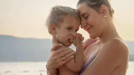 Foto de Silueta de madre abrazando al bebé hijo en la playa del mar al atardecer. Vacaciones, vacaciones de verano y turismo. - Imagen libre de derechos