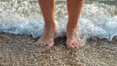 Foto de Las olas del mar rodando y rompiendo en los pies femeninos de pie en la playa del océano. Vacaciones, vacaciones de verano y turismo. - Imagen libre de derechos