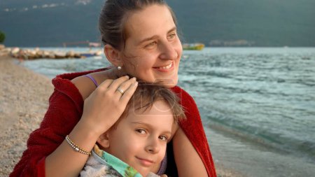 Téléchargez les photos : Jeune mère aimante embrassant son petit fils couvrant de serviette de plage et se sentant froid en mer. Vacances, vacances d'été et tourisme. - en image libre de droit