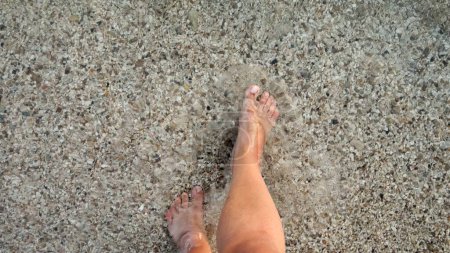 Téléchargez les photos : Pieds masculins marchant sur la plage de sable de l'océan et des cailloux à travers l'eau de mer calme et claire. - en image libre de droit