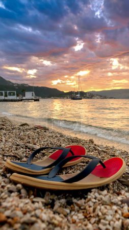 Photo for Vertical shot of beach flip-flops lying on the sand against sunset sky and rolling sea waves. Concept of summertime, vacation and beach holidays - Royalty Free Image
