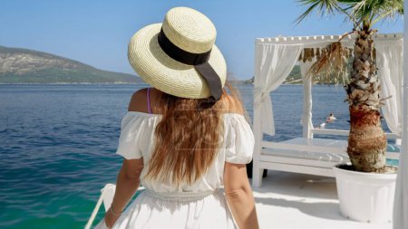 Téléchargez les photos : Vue arrière de la femme élégante en chapeau et robe longue se détendre en marchant sur la jetée de la mer en bois par une journée d'été ensoleillée. - en image libre de droit