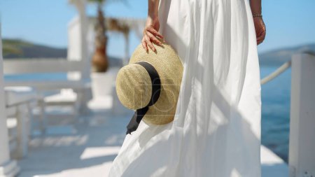 Photo for Closeup of woman in long white dress holding summer straw hat and walking on white wooden pier. - Royalty Free Image