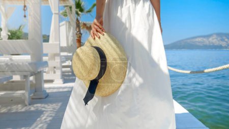 Téléchargez les photos : Dame en robe légère et chapeau de paille marchant sur une jetée en bois, rayonnant vacances, voyage et été - en image libre de droit
