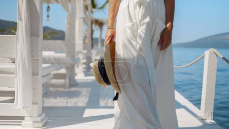 Photo for Closeup of elegant woman holding summer straw hat walking on wooden pier at the sea beach on sunny summer day. - Royalty Free Image